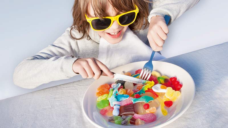 Criança de óculos escuros com face a garfo, sentada à frente de um prato cheio de gomas - alusão a aditivos nestas guloseimas